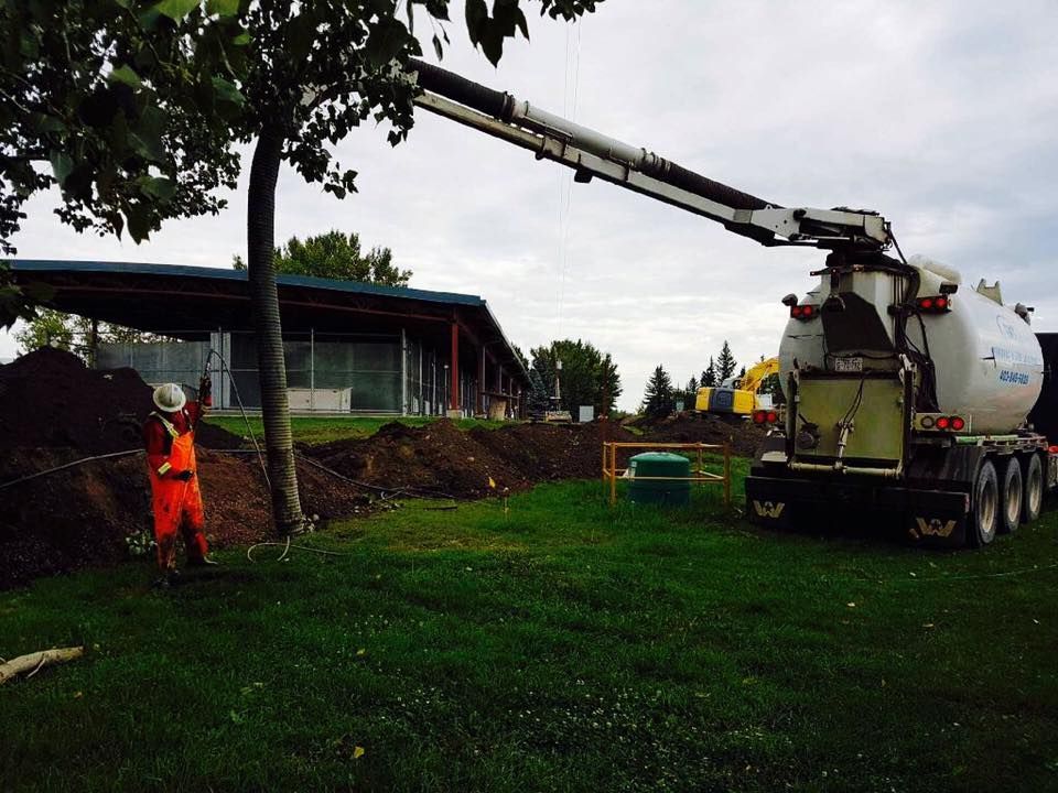 A construction worker in high-visibility gear operates a vacuum truck on a grassy site with piles of excavated soil.