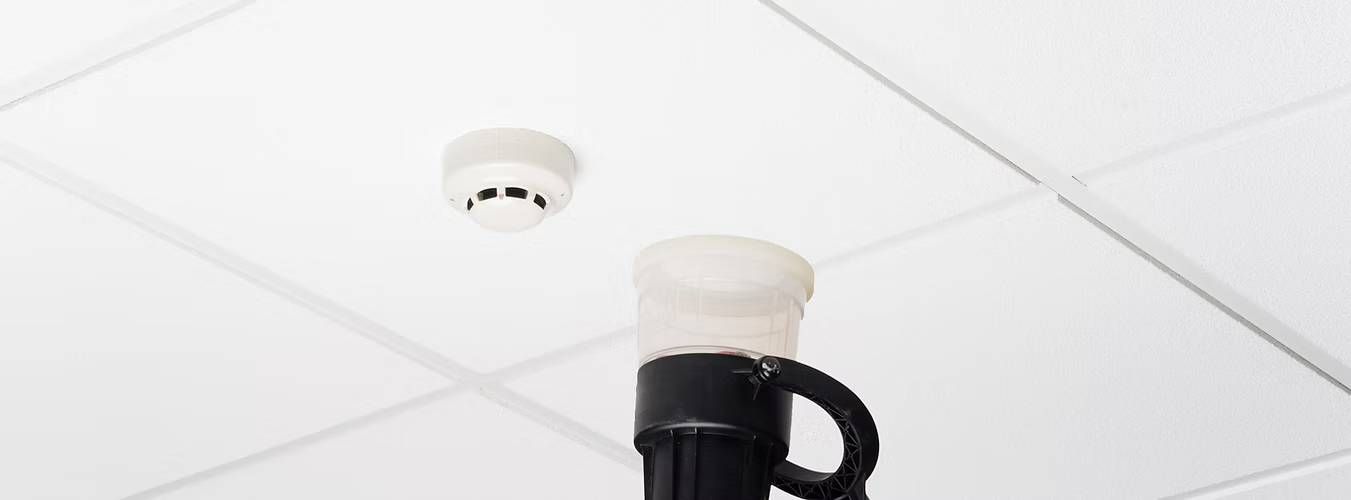A smoke detector is mounted on a white tiled ceiling
