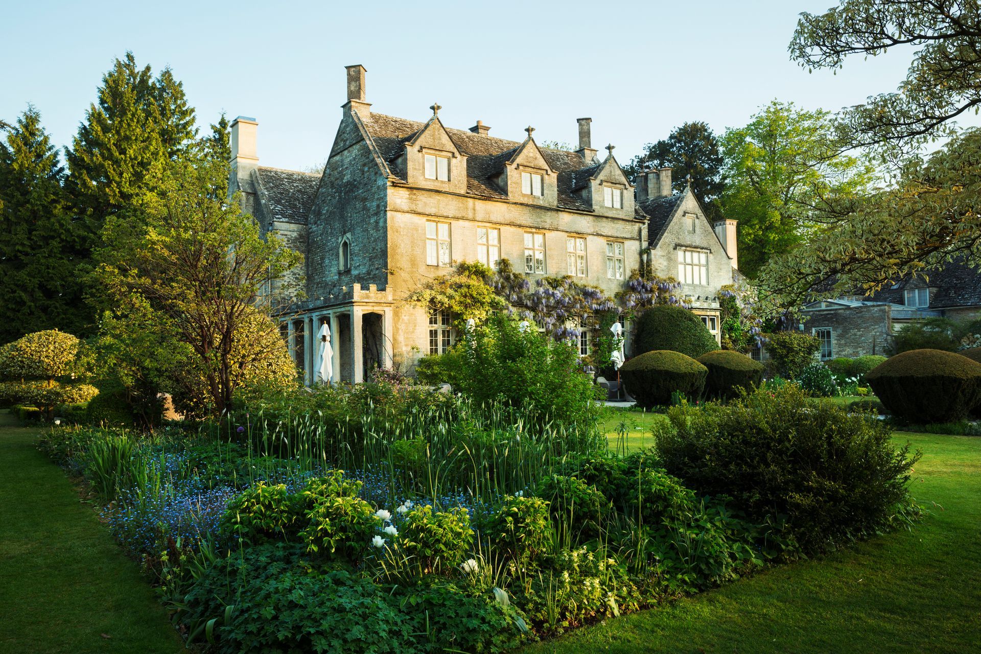 Large stone manor house with garden and lush greenery. Sunlight illuminates the building and surrounding plants.