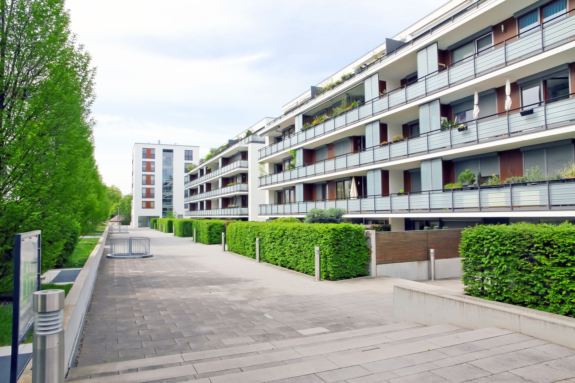 Apartment buildings with balconies, pathway lined with green hedges and trees.