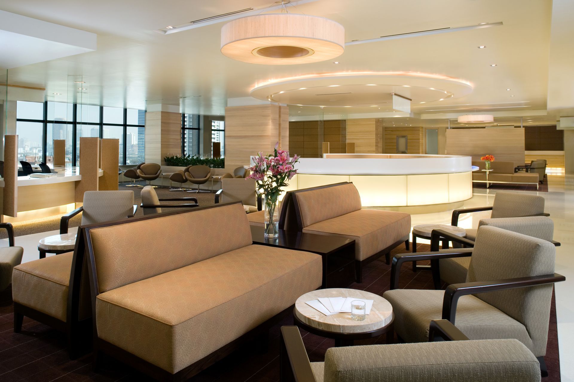 Lounge area with tan sofas and chairs, small tables, and a curved reception desk. Soft lighting, neutral tones.