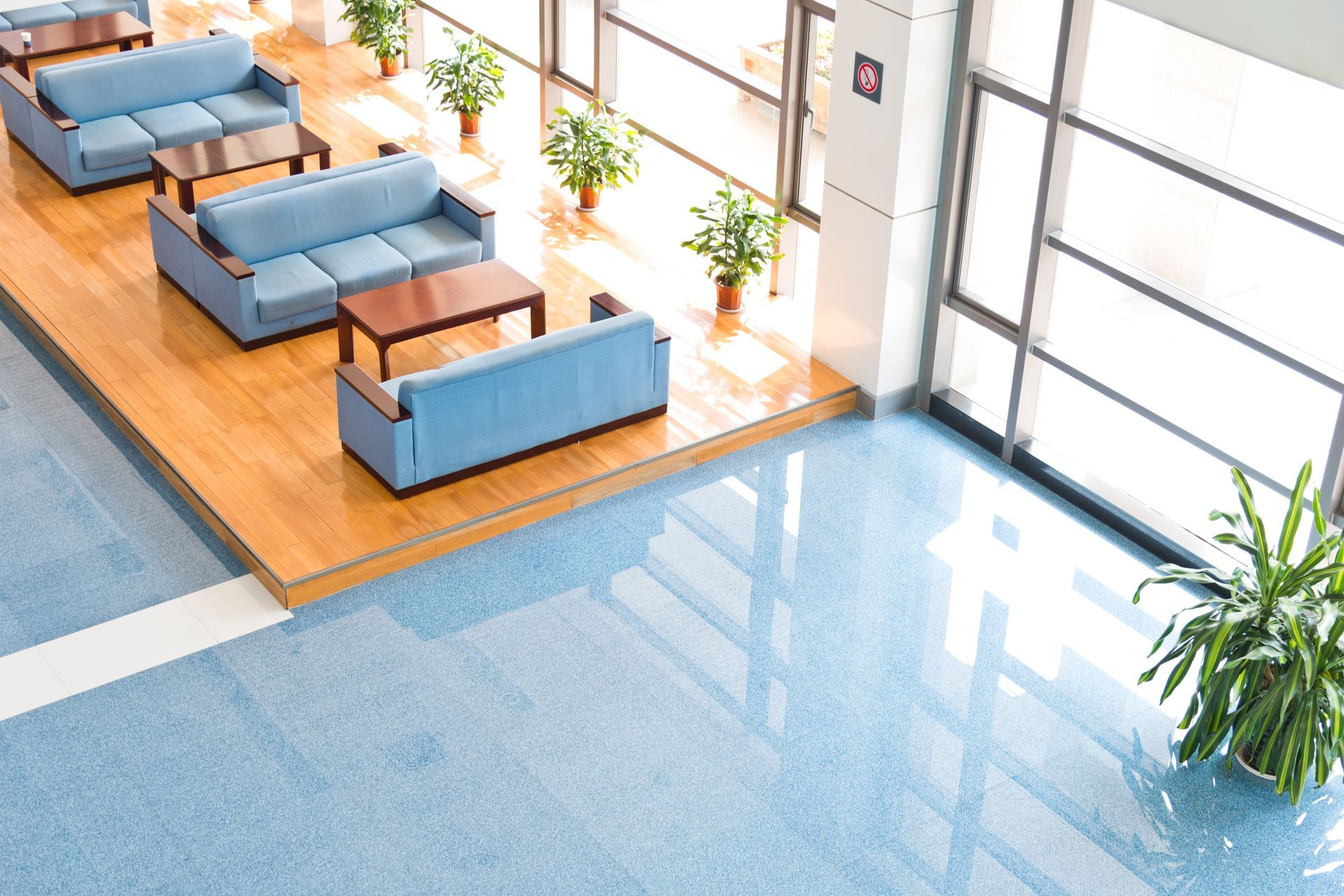 Bright waiting area with blue sofas and wooden tables on a raised platform, next to a large window.