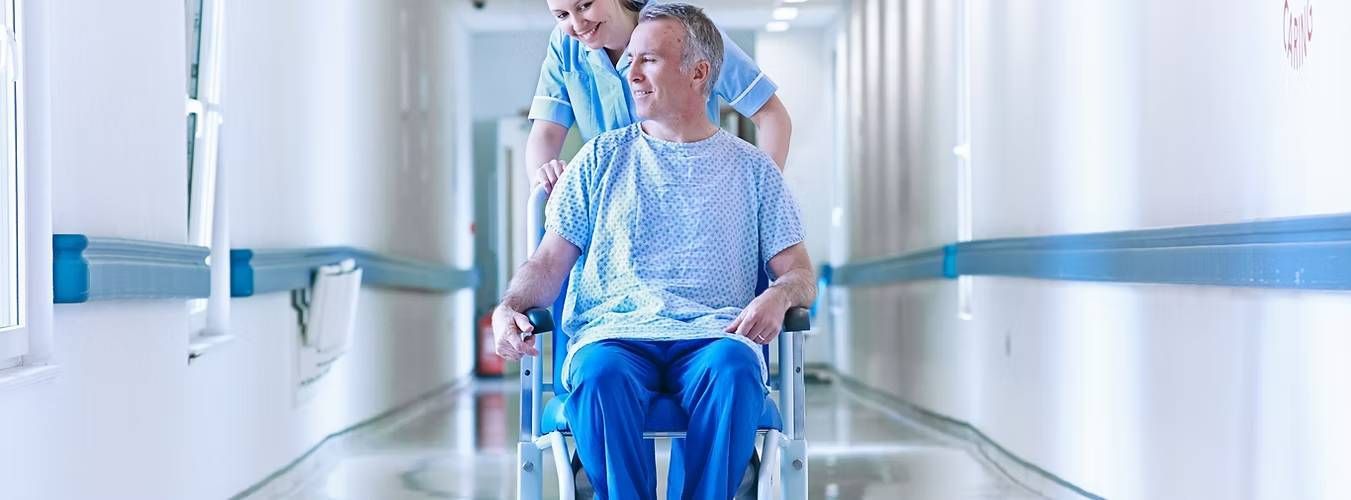 A nurse pushes a smiling man in a wheelchair down a bright hospital corridor
