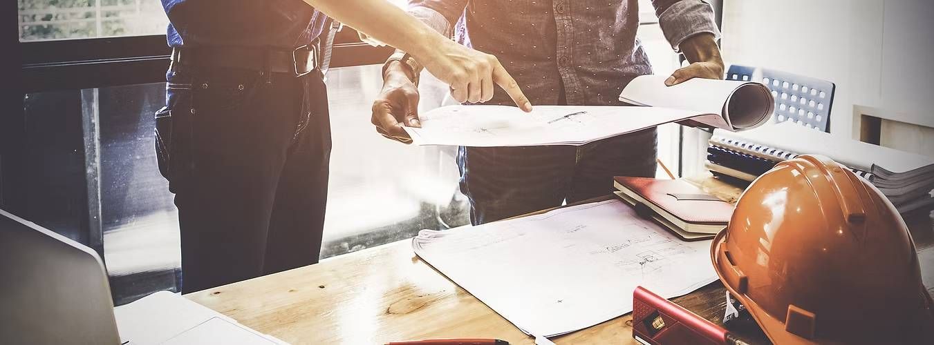 Two people examine blueprints on a wooden table
