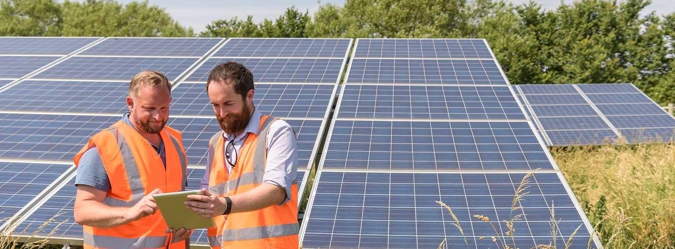 Two men in orange safety vests examine a tablet in front of large solar panels
