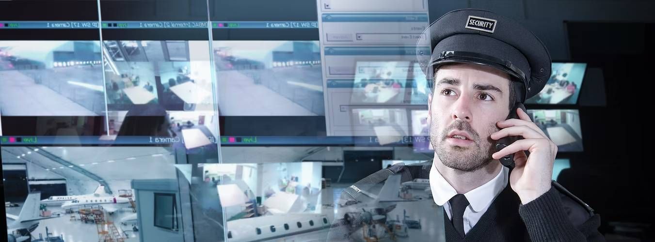 A security guard in uniform talks on a phone, surrounded by multiple surveillance screens