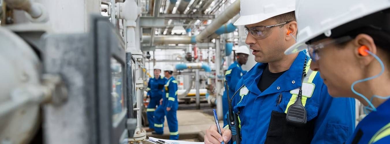 Workers in blue safety gear and helmets inspect industrial pipes, with one taking notes
