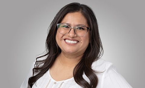Smiling person with glasses and long dark hair in a white top against a gray background