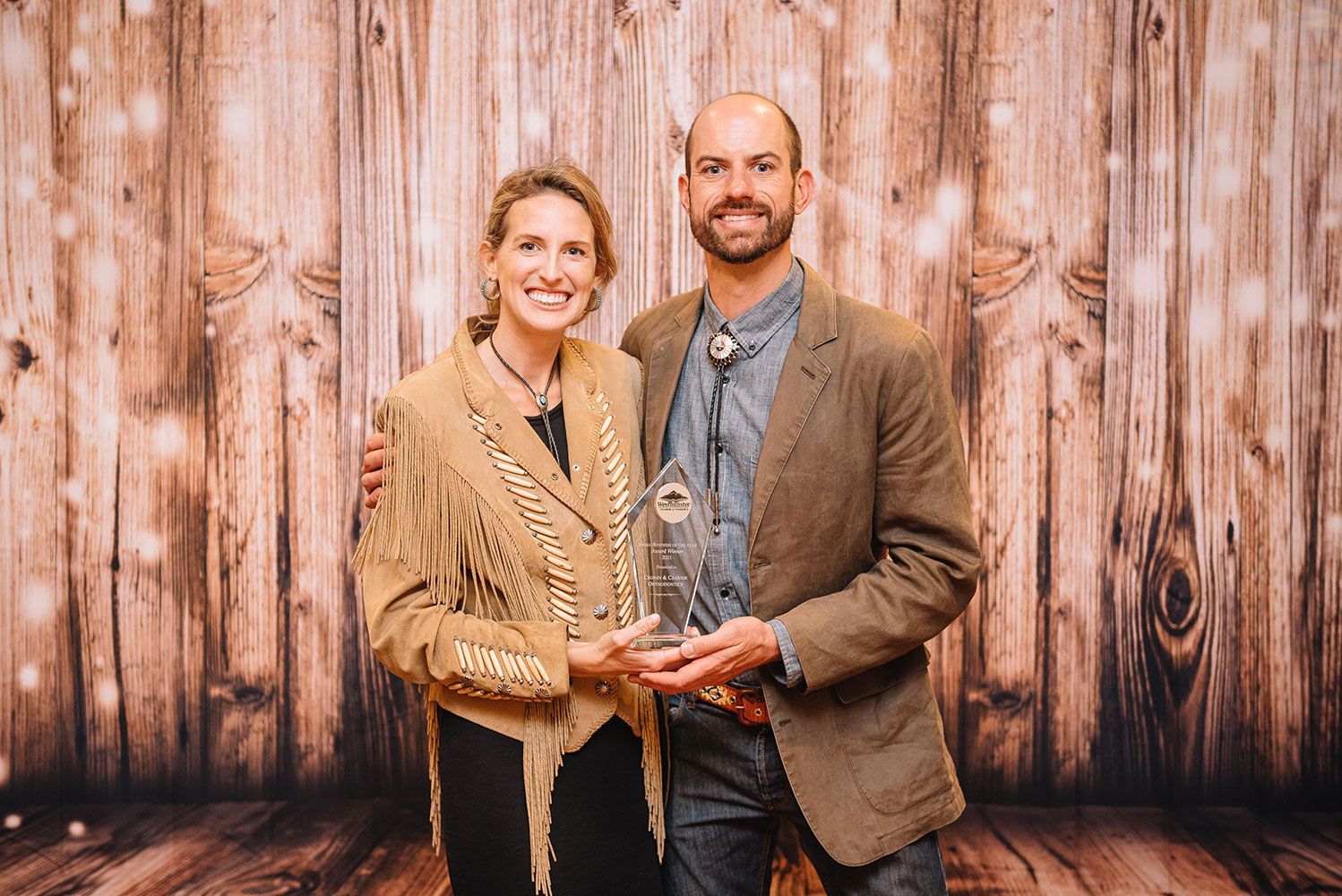 Two people holding a clear award in front of a rustic wood wall, smiling at the camera