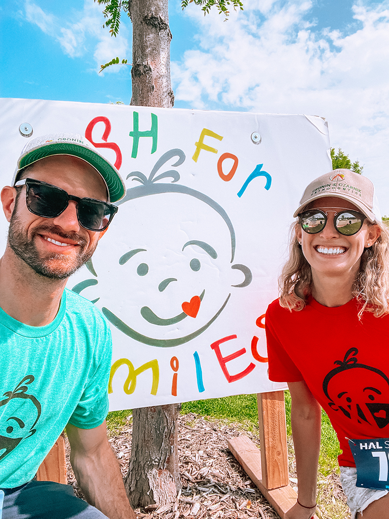 Two smiling people pose by a colorful hand-drawn sign on a beach with palm trees.