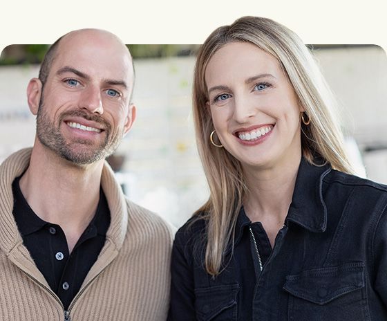 Two smiling people standing outdoors, one in a beige sweater and one in a black shirt and jacket.