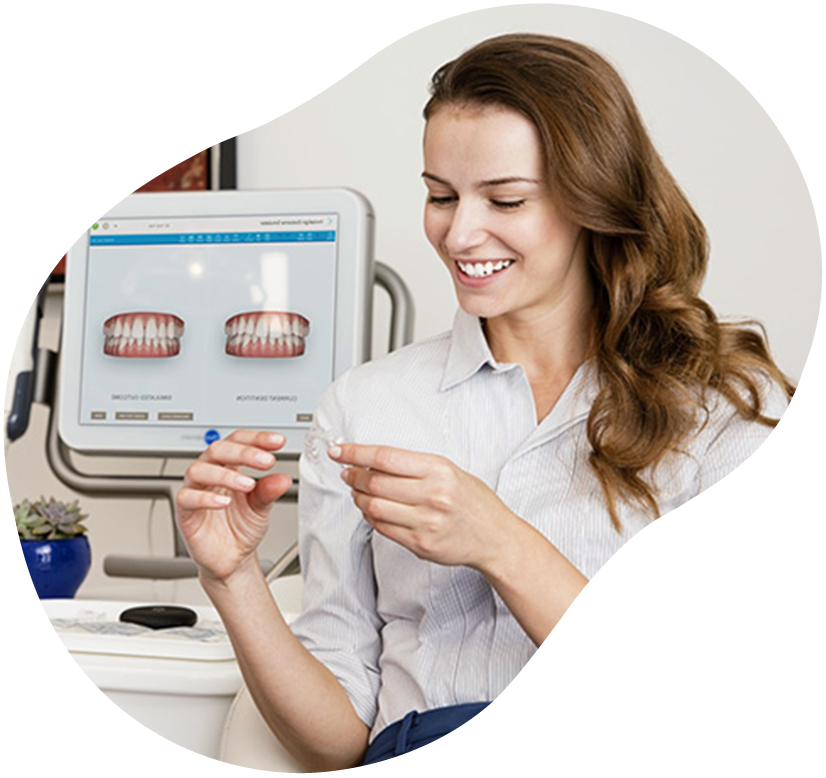 Woman smiling at a dental office, holding a dental aligner beside a monitor showing teeth models