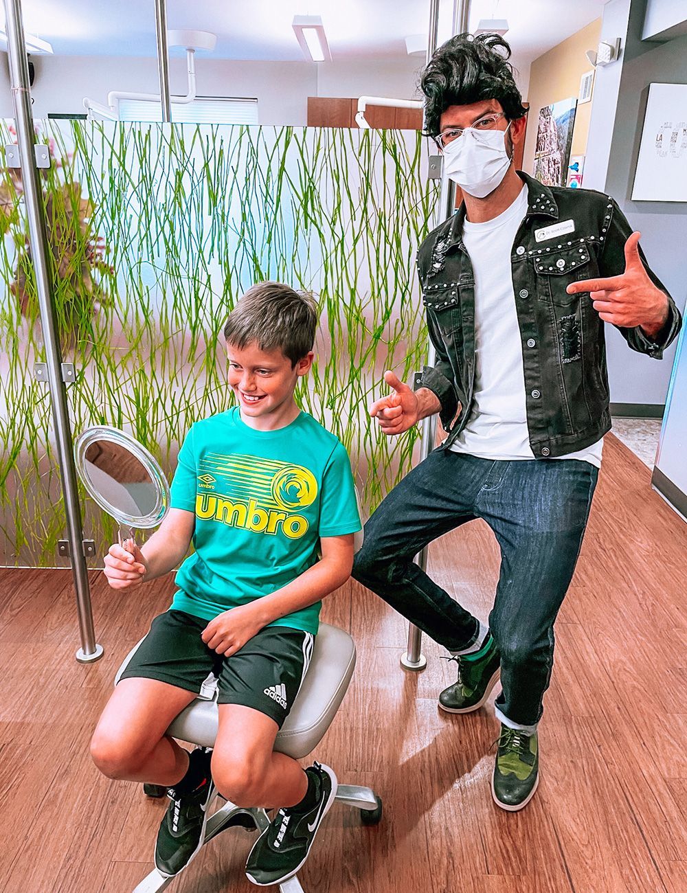 Two people in a clinic hallway, one seated and one standing, both wearing masks and giving thumbs up.