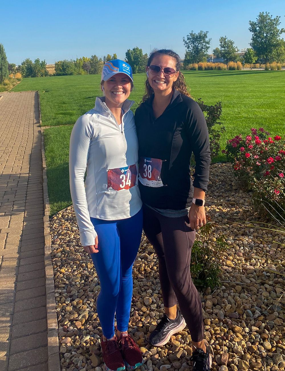 Two runners posing outdoors on a sunny path, wearing race bibs and athletic clothes.