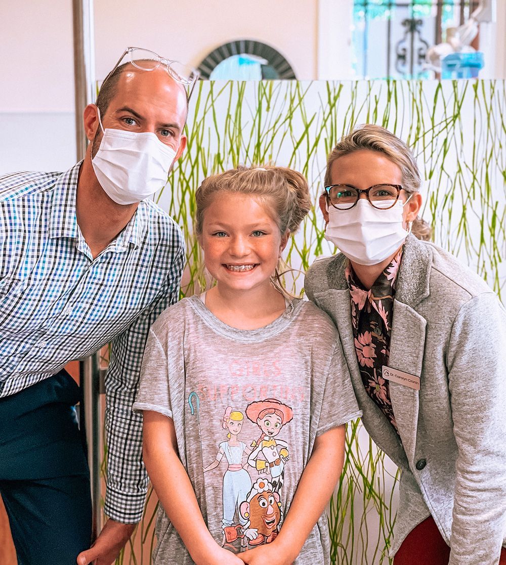 Three masked people smiling indoors, standing in front of tall green plants.