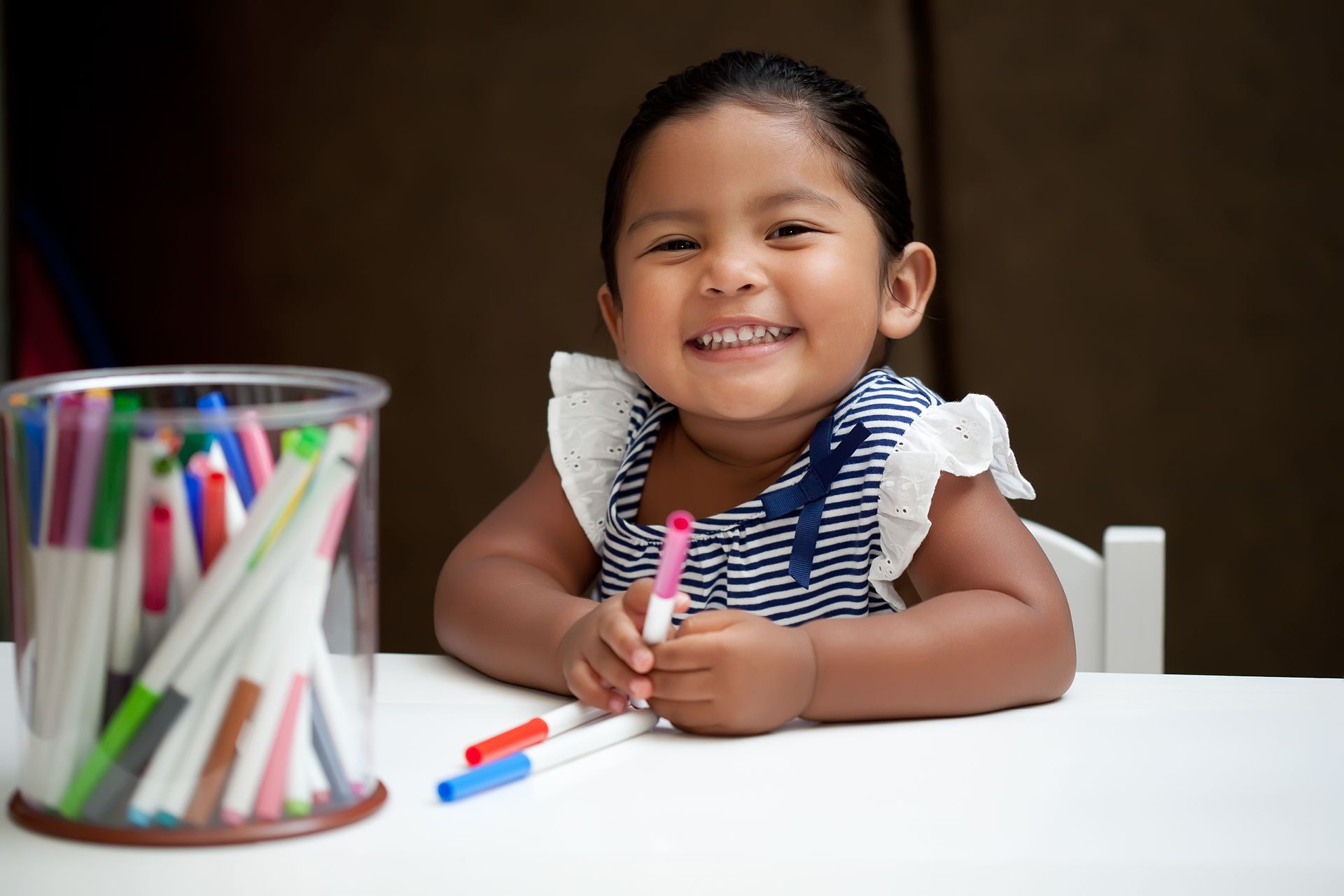 Une petite fille est assise à une table, tenant un marqueur et souriant.