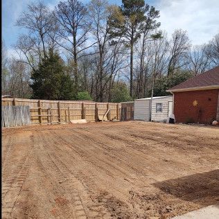 Dirt yard with wooden fence, shed, brick building, and bare trees under a cloudy sky.