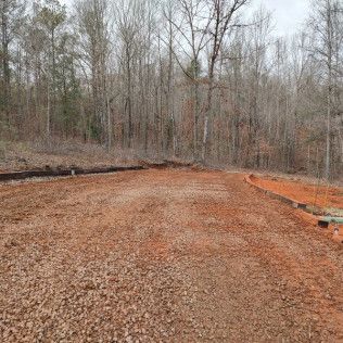 Gravel road in a wooded area, trees in the background. Red dirt and gravel in the foreground.
