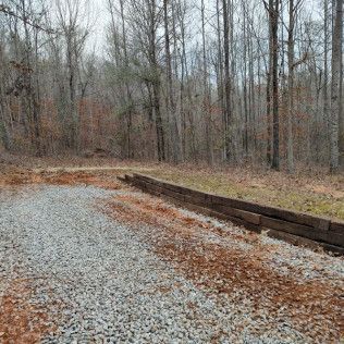 Gravel driveway with a wooden border, leading into a wooded area with bare trees.