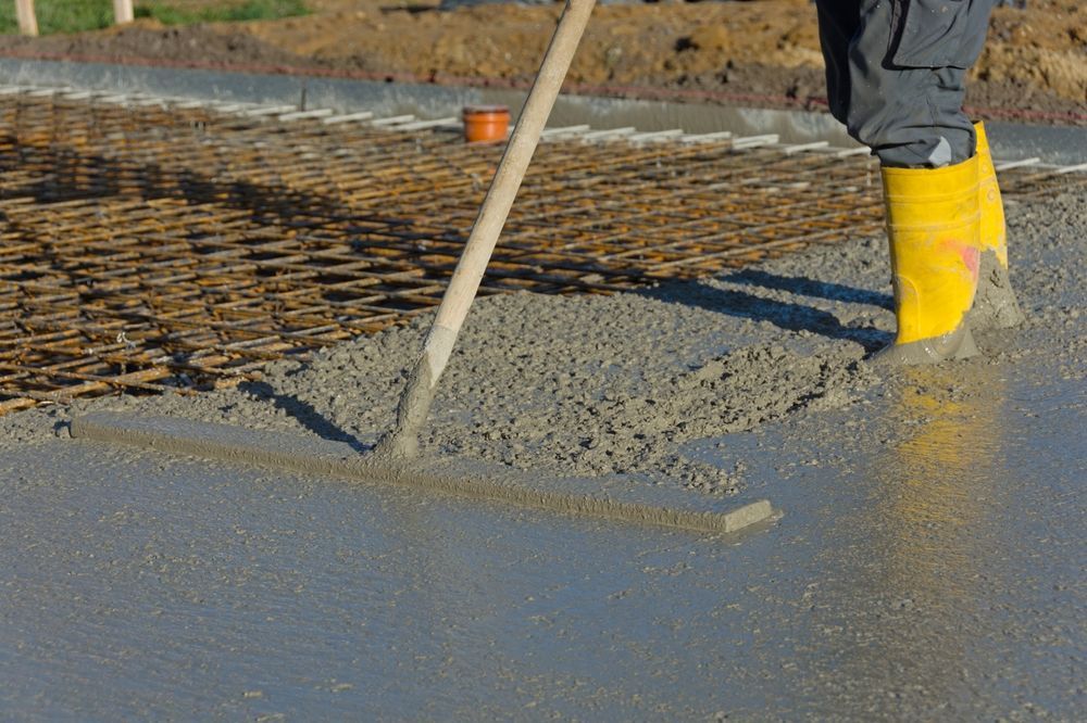Construction worker rakes wet cement, leveling a surface; yellow boots, steel reinforcement, outdoors.