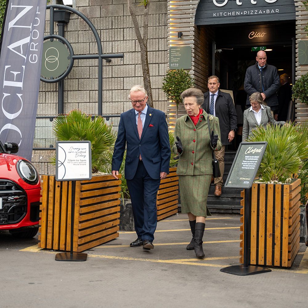 Princess Anne and a man in a suit exit a restaurant, followed by others. A red Mini Cooper and sign are visible.