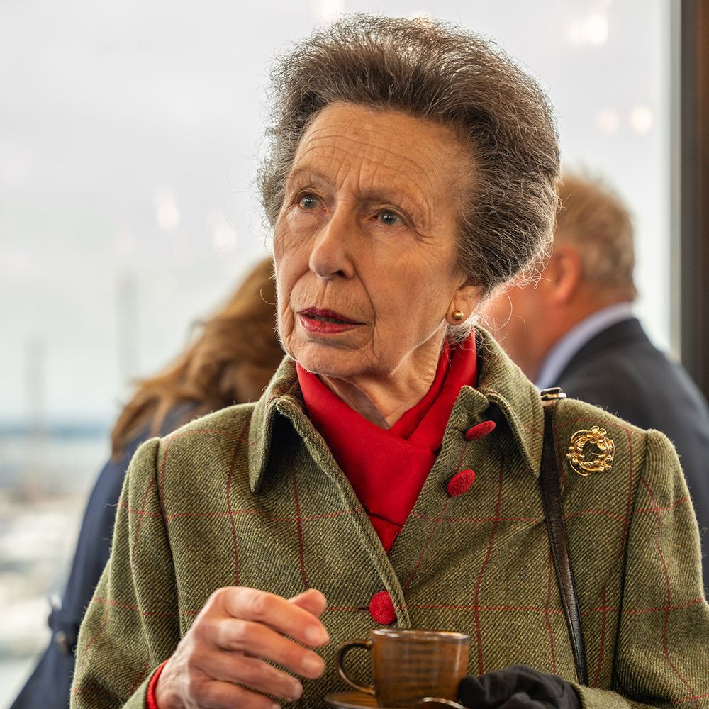 Princess Anne in a green tweed jacket, red scarf, and holding a teacup, looking to her right.