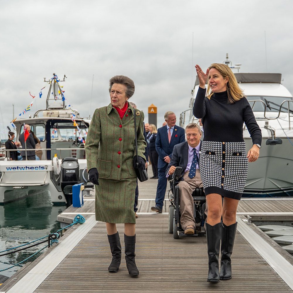 Princess Anne and a woman walk along a dock, waving. Man in wheelchair follows.