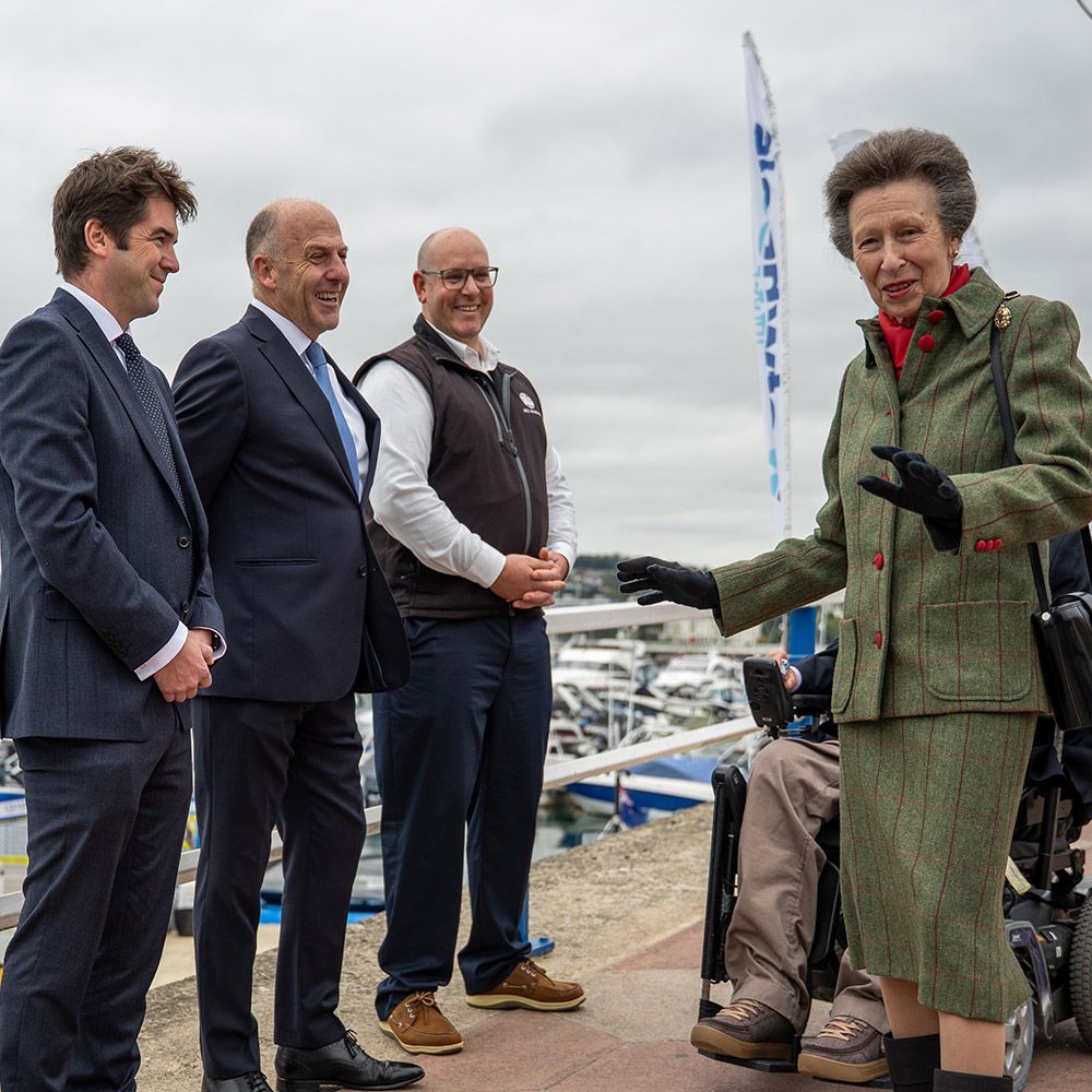 Princess Anne speaks outdoors to three men near a marina, gesturing with her hands.