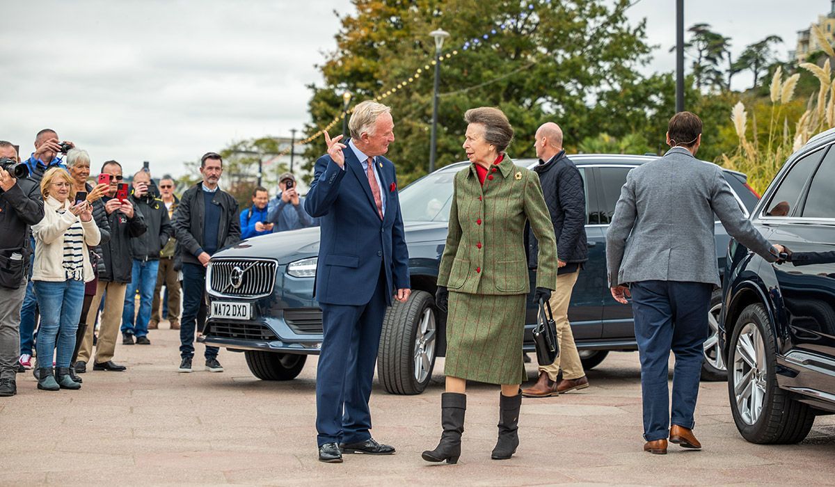 Princess Anne and a man in a suit stand near dark SUVs, waving to a crowd outdoors.