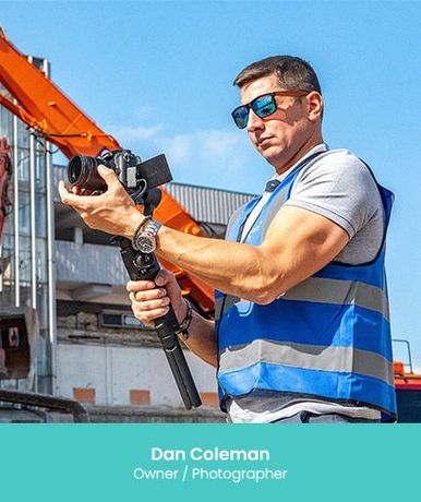 Man holding a camera, wearing sunglasses and a reflective vest, standing near construction equipment.