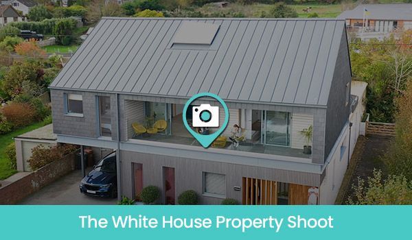 Modern gray house with car parked beneath a balcony. Greenery surrounds, and a camera icon is overlaid.