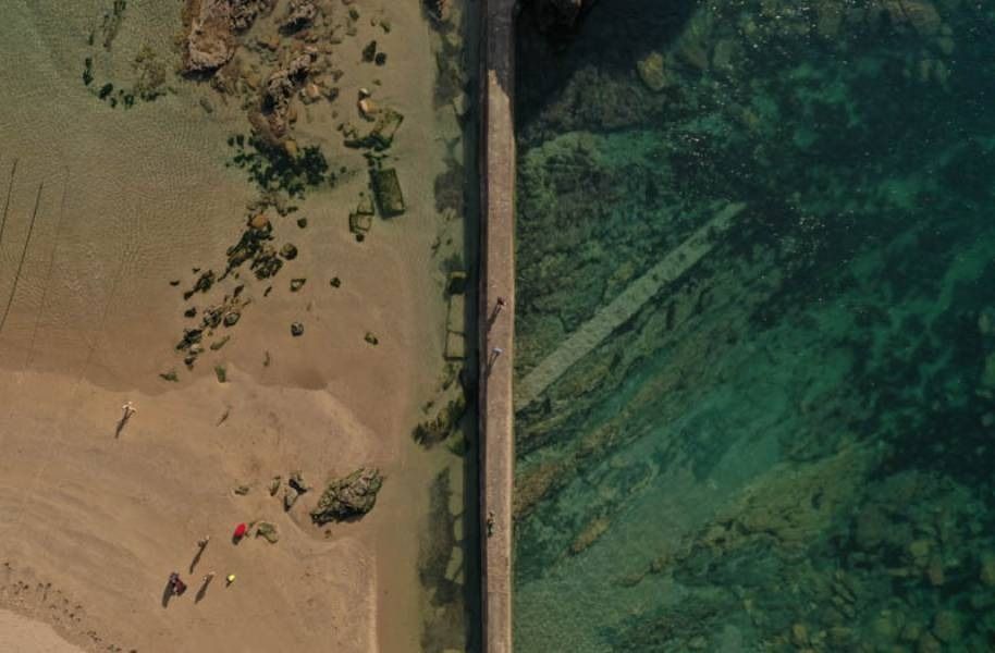 Aerial view of a long, narrow pier dividing sandy beach and turquoise water. People walking on the pier and beach.
