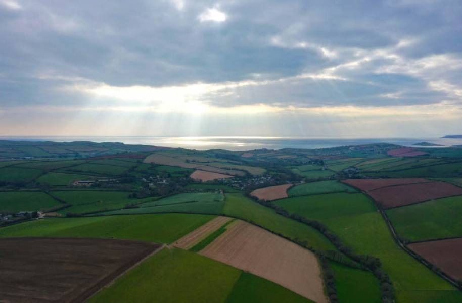 Aerial view of patchwork fields and a coastal landscape, with a bright sky and ocean in the distance.
