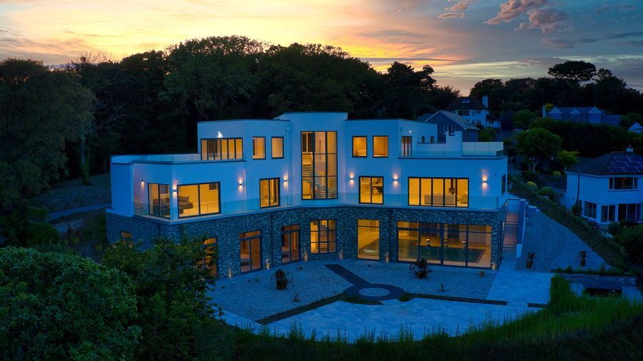 Modern white and stone house at dusk, lit interior, surrounded by trees.
