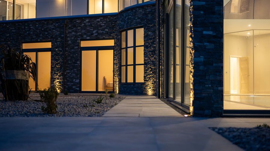 Exterior of a modern building at dusk, with stone and glass walls, lit walkway.