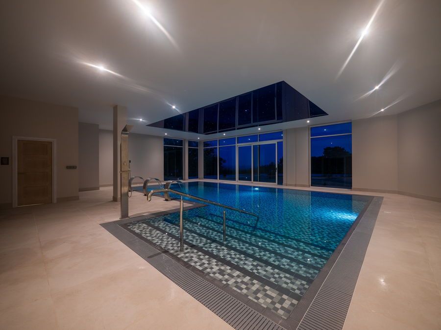 Indoor pool with glass walls, blue water, and overhead lighting. The room is light with beige flooring.