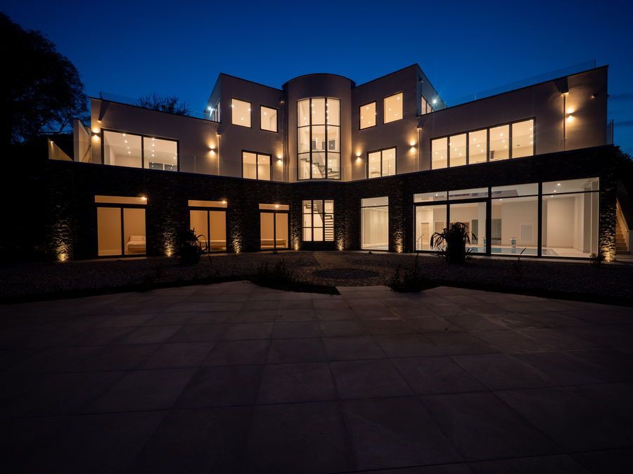 Modern three-story house at dusk, lit interior lights, glass windows, curved center section, dark stone exterior, large patio.