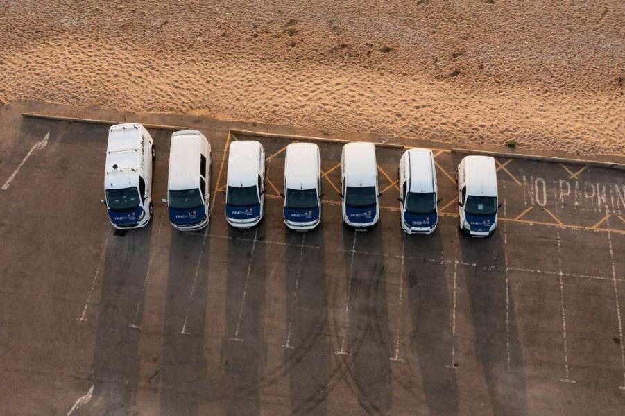 Seven white vans parked in a row on asphalt near a beach, a “no parking” marking on the side.