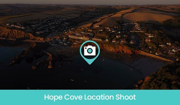 Aerial view of Hope Cove, Devon, with a camera icon overlaid. Coastal town and beach at sunset.