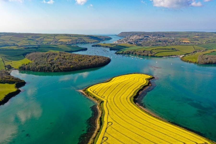 Aerial view of a bright blue river flowing through green hills and a yellow rapeseed field, reaching the ocean.