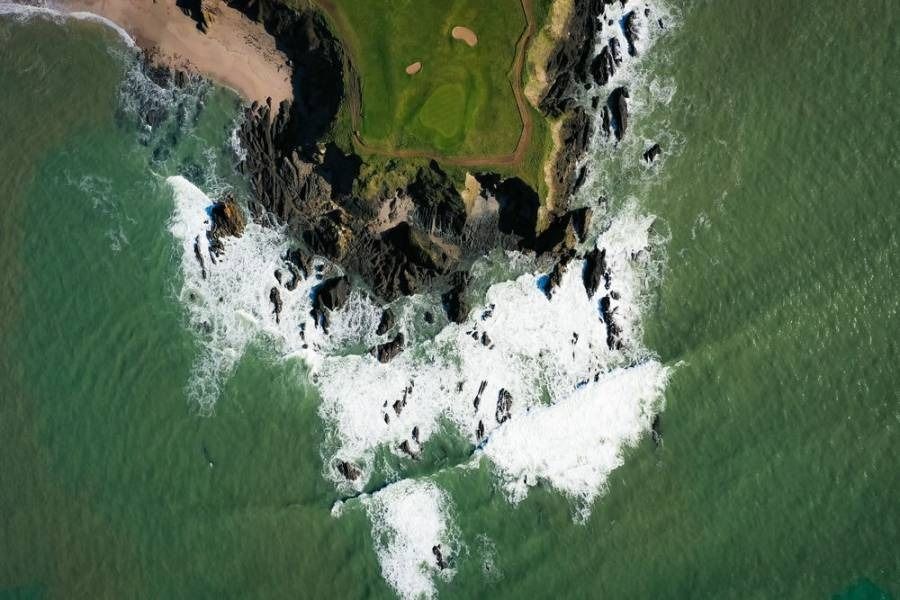 Aerial view of rocky coastline with waves crashing, adjacent to a green golf course.