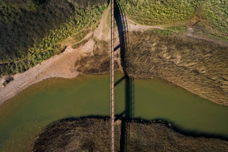 Overhead view of a bridge crossing a river with grassy banks; bridge casts a long shadow.