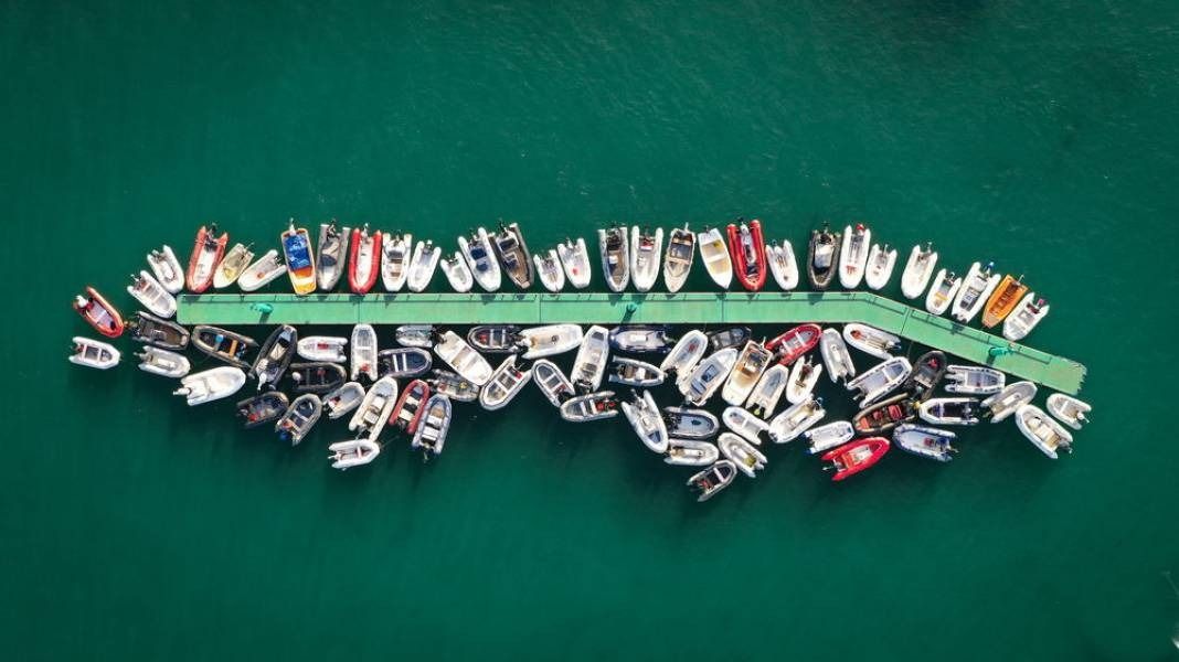 Overhead view of many small boats clustered around a green pier in turquoise water.