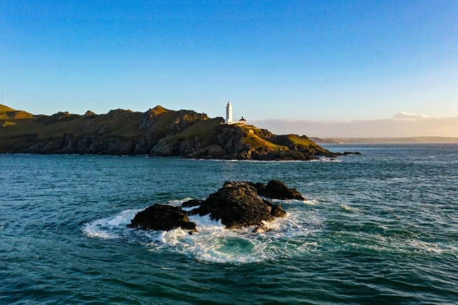 Ocean waves crashing on rocks, a lighthouse atop a cliff under a blue sky.