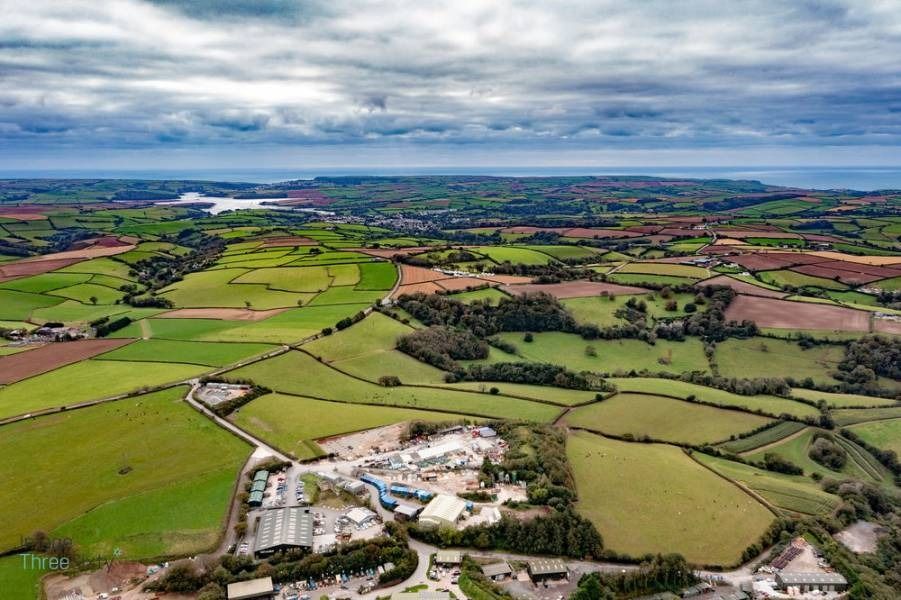 Aerial view of green fields, trees, and buildings under a cloudy sky, with ocean in the distance.