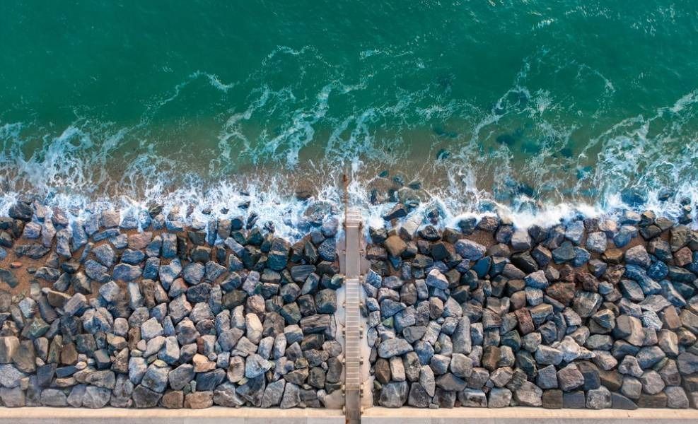 Ocean waves crashing against a rocky stone barrier. A small walkway extends into the sea.