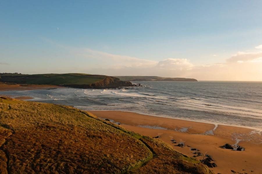 Beach with golden sand, waves, cliffs, and a blue sky at sunset.