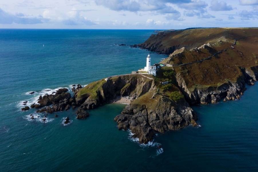 White lighthouse on a rocky peninsula, surrounded by blue ocean under a cloudy sky.