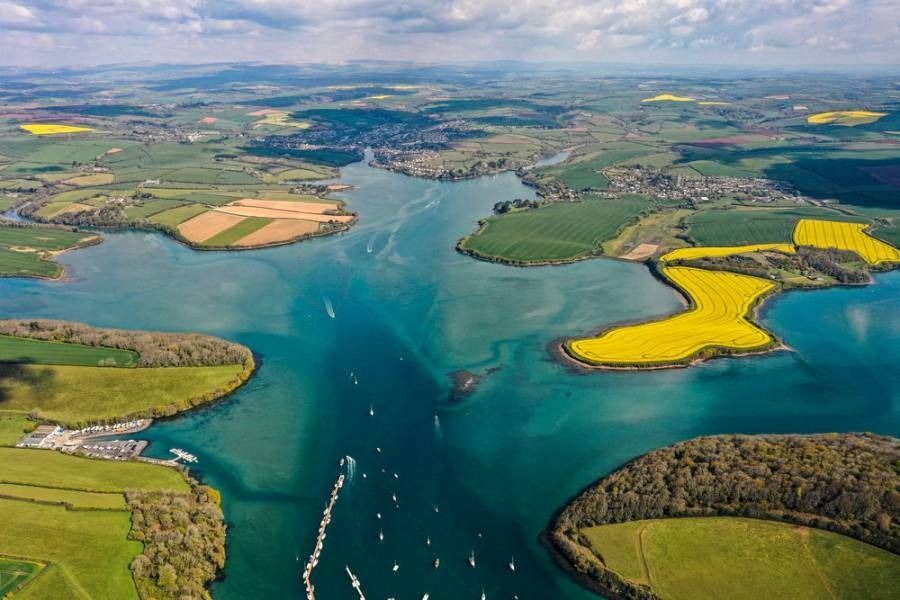 Aerial view of a teal-colored river winding through green fields and yellow rapeseed blooms. Boats dot the water.