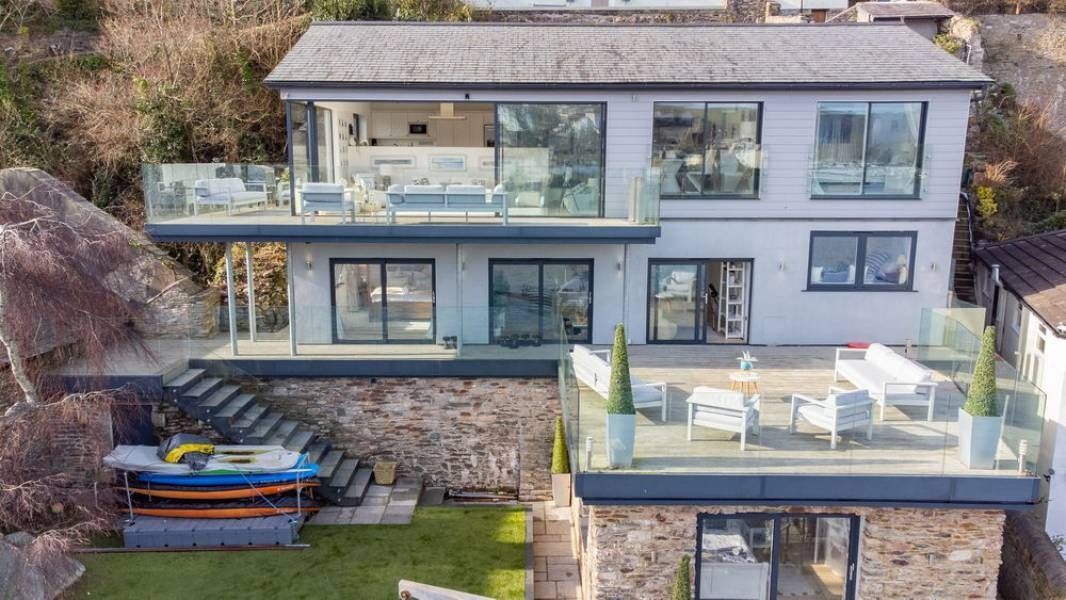 Modern two-story house with glass balconies, overlooking water. Gray exterior, stone base. Stairs lead down to a boat rack.